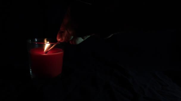 Burning red candle with flickering flame, hand lighting a candle with a match on dark background with copy space — Stock Video