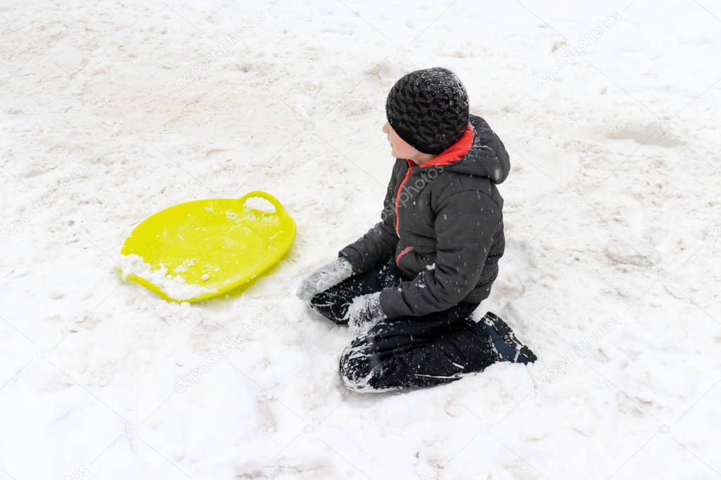 A boy of seven years old sitting on the snow and a green plastic saucer sled lying near him. Concept of winter activities, recreation and children's entertainment.