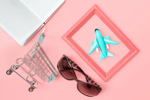 Shopping cart, notebook, plane and sunglasses on pink background, flat lay - shopping tourism concept.