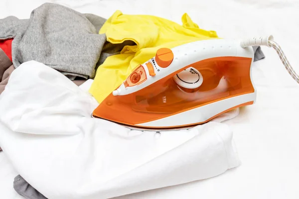 Оранжевый современный электрический утюг и куча неглаженной одежды вблизи — стоковое фото