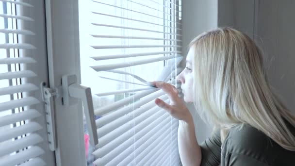 Любопытная молодая женщина шпионит, подглядывает через жалюзи в своем доме — стоковое видео