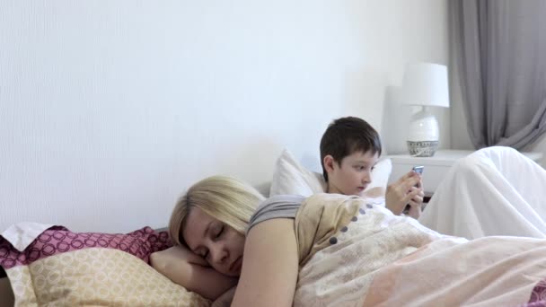 疲れた女性の母親が寝て、彼女の太陽がベッドに座って、ガジェット、スマートフォンでモバイルゲームをプレイした後に目覚める。注意の欠如と子供の孤独の概念 — ストック動画