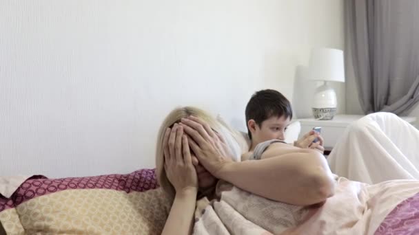 Κουρασμένη γυναίκα μητέρα ξυπνάει μετά τον ύπνο και τον ήλιο της κάθεται στο κρεβάτι και παίζοντας κινητά παιχνίδια σε gadget, smartphone. Έλλειψη προσοχής και της έννοιας της μοναξιάς των παιδιών — Αρχείο Βίντεο
