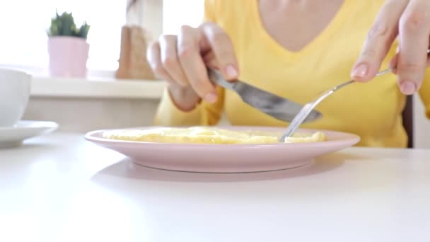 Mujer sentada junto a la mesa, comiendo un omlet con un folklore y un cuchillo y tomando café o té para desayunar en casa o restaurante — Vídeo de stock