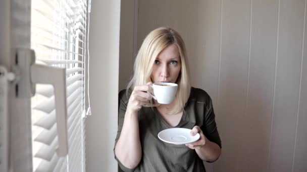 年轻的悲伤的想法美丽的金发碧眼的女人站在窗边, 早上百叶窗, 喝咖啡 — 图库视频影像