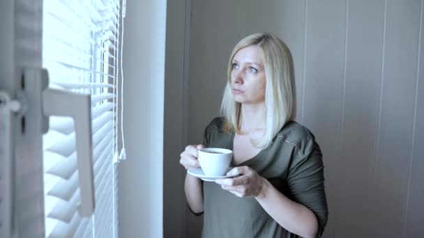 Junge traurig denkende schöne blonde Frau, die morgens mit Jalousien am Fenster steht und Kaffee trinkt — Stockvideo