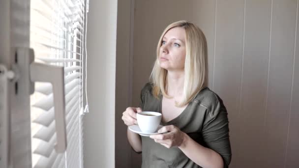 Молодая грустная, проницательная, красивая блондинка, стоящая у окна с жалюзи по утрам и пьющая кофе — стоковое видео