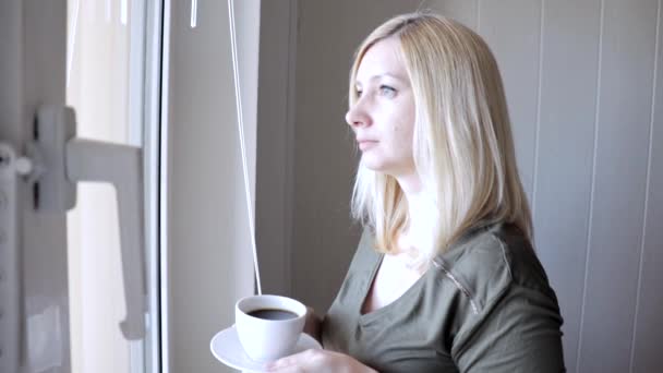 Junge traurig denkende schöne blonde Frau, die morgens mit Jalousien am Fenster steht und Kaffee trinkt — Stockvideo