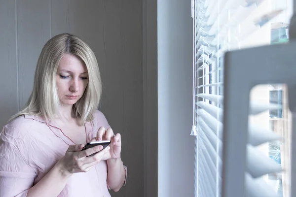 Сумна апатична самотня жінка, що стоїть біля вікна і смс-повідомлення в смартфоні вдома або в готелі, розлучення, депресія і концепція апатії — стокове фото