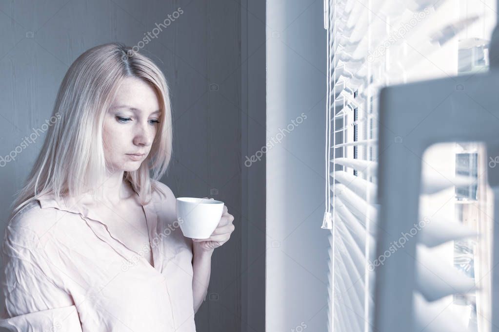 Sad apathetic lonely woman with a cup of coffee looking through a window at home or hotel, divorce, depression and apathy concept