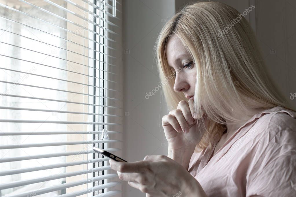 Sad apathetic lonely woman standing near the window and texting messages in smartphone at home or hotel, divorce, depression and apathy concept