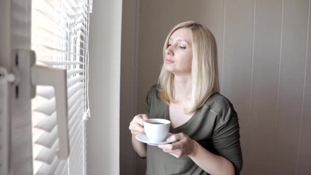 Junge traurig denkende schöne blonde Frau, die morgens mit Jalousien am Fenster steht und Kaffee trinkt — Stockvideo