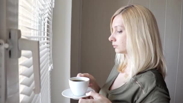 Jeune fille triste pensant belle femme blonde debout près de la fenêtre avec des stores le matin et boire du café — Video