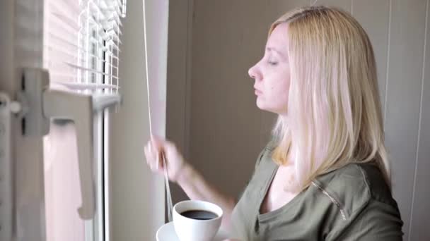 Junge traurig denkende schöne blonde Frau, die morgens am Fenster steht, Kaffee trinkt und Jalousien hochzieht — Stockvideo