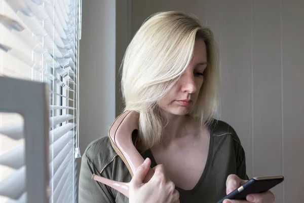 一个白人金发女郎站在窗口附近，穿着高跟鞋，用她的手机上网冲浪 - 网上购物概念 — 图库照片