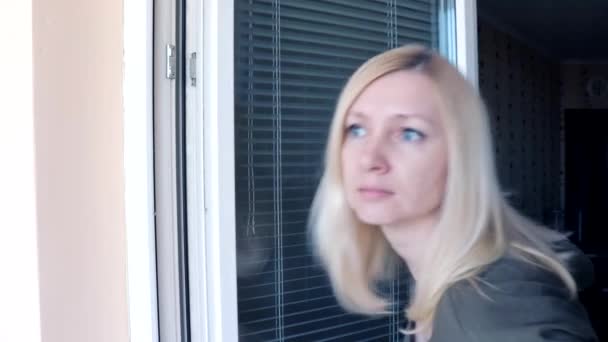 Молода приваблива домогосподарка, блондинка, що стоїть біля відкритого вікна, дивиться на когось, махає рукою і вигукує привіт, вітає когось — стокове відео