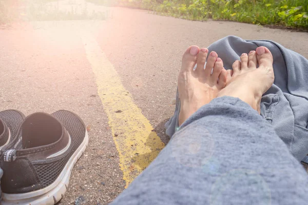 Žena s bosými nohama a batohu, která sedí na asfaltové silnici se žlutou jednoduchou silniční linkou čekající na stopku, letní sezonu — Stock fotografie