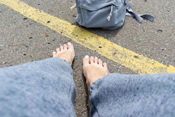 Donna a piedi nudi e zaino seduta su una strada asfaltata con una sola linea stradale gialla in attesa del veicolo per l'autostop, stagione estiva — Foto Stock