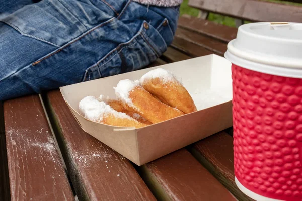 Closeup of beignet pastries, donuts in a paper takeaway box and a cup of coffee on a bench in park. The delicious deserts covered in powdered sugar, bought from a street fast food
