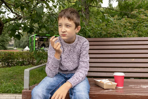 Мальчик-подросток, сидящий на скамейке в парке и поедающий выпечку, пончики и пьющий кофе или чай из коробки для еды на вынос и чашки, концепция уличного фаст-фуда — стоковое фото