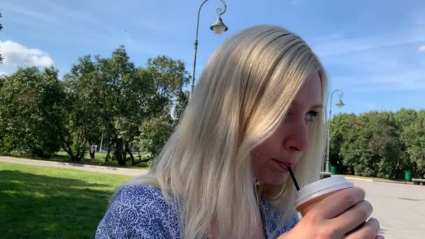 Νεαρά ξανθιά καυκάσια γυναίκα κάθεται στον πάγκο στο καλοκαιρινό πάρκο της πόλης και πίνοντας καφέ χρησιμοποιώντας ένα άχυρο από ένα πακέτο χαρτιού μίας χρήσης — Αρχείο Βίντεο