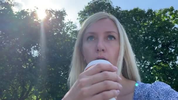 여름 도시 공원에서 벤치에 앉아 일회용 종이 테이크 아웃 컵에서 빨대를 사용하여 커피를 마시는 젊은 금발 의 백인 여성 — 비디오