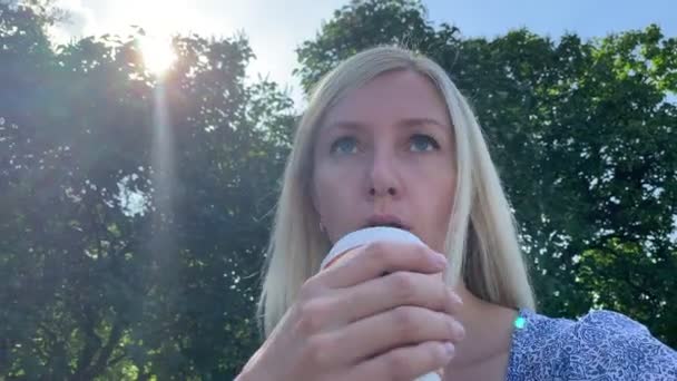 Jeune femme caucasienne blonde assise sur le banc dans le parc de la ville d'été et buvant du café à l'aide d'une paille d'une tasse à emporter en papier jetable — Video