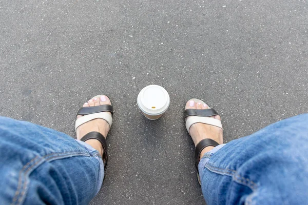 Una taza de papel desechable kraft para llevar con taza blanca en el camino de asfalto entre las piernas de la mujer pies con sandalias de cuero y jeans, descanso de café de verano en el parque de la ciudad, espacio para copiar —  Fotos de Stock