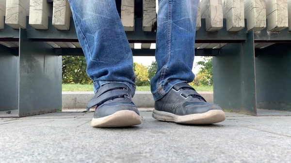 Piernas pies de un adolescente aburrido sentado en un banco en el parque urbano de la ciudad y con jeans y zapatos —  Fotos de Stock
