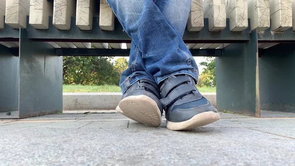 Gambe piedi di un adolescente annoiato seduto su una panchina nel parco urbano della città e indossando jeans e scarpe, è pigro e incrociando le gambe — Foto Stock