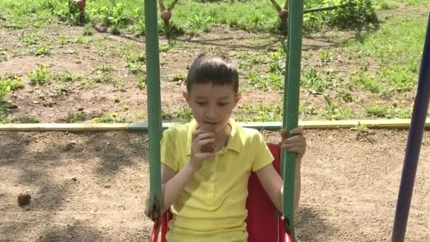 Preteen kaukaski chłopiec siedzi na huśtawce i jedzenia smaczne pyszne lody na placu zabaw dla dzieci w okresie letnim — Wideo stockowe