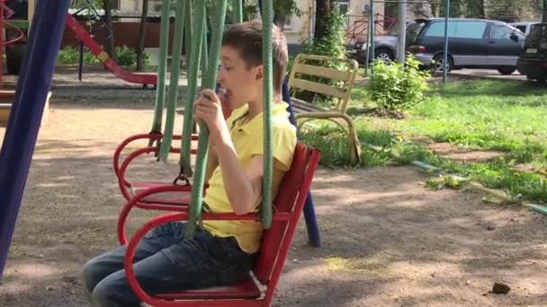 Ένα προεφηβικό καυκάσιο αγόρι που κάθεται σε μια κούνια και τρώει νόστιμο νόστιμο παγωτό στην παιδική χαρά του καλοκαιριού — Αρχείο Βίντεο