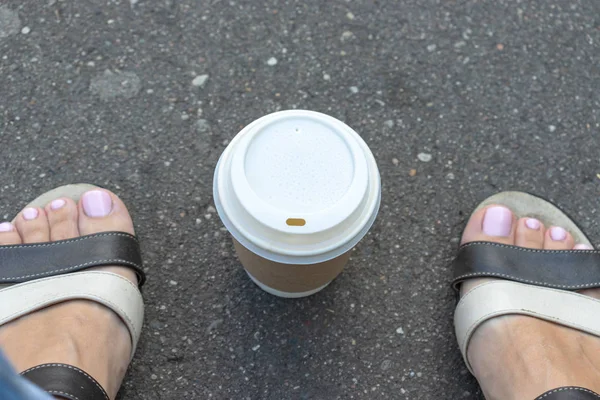 Una taza de papel desechable kraft para llevar con taza blanca en el camino de asfalto entre las piernas de la mujer pies con sandalias de cuero, descanso de café de verano en el parque de la ciudad —  Fotos de Stock