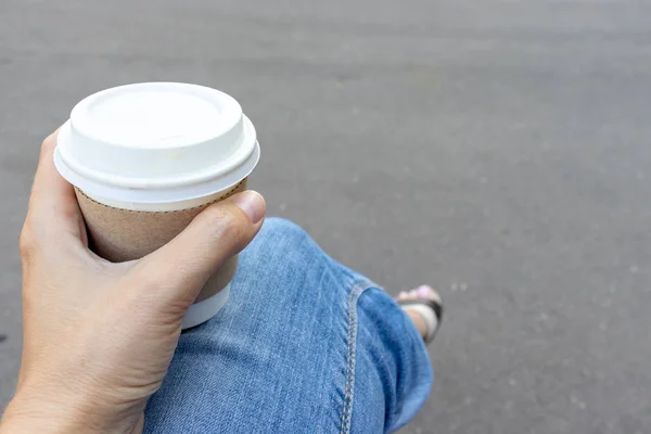 Woman sitting on a bench outdoor and holding a kraft disposadle paper cup of coffee with a white cup in hand — Stock Photo, Image