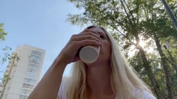 Jovem loira sentada no banco no parque da cidade de verão e beber café descartável copo de papel takeaway com um copo de forma de rosto humano preto — Vídeo de Stock