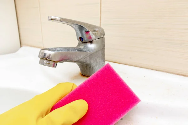 Рука в жёлтых резиновых перчатках с губкой для мытья грязного крана с лимескалем, кальцинированный водопроводный кран с лаймовой шкалой на умывальнике в ванной комнате, концепция уборки дома — стоковое фото