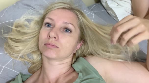 Задумчивая, расслабленная блондинка лежит в постели и трогает, гладит волосы — стоковое видео