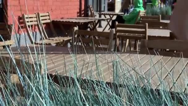 Straat cafe met lege houten tafels en wazig mensen op de achtergrond, decoratief gras bewegen in de wind op de voorgrond — Stockvideo