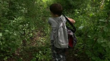 Orman yolunda yürüyen sırt çantasıyla gezen genç bir çocuk. Etrafında bir sürü yüksek çim ve orman var. Bir sopayla çimleri ve çalıları deviriyor.