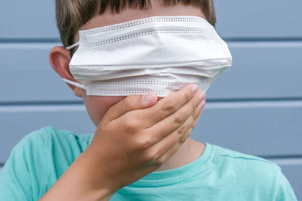 Μια αστεία άποψη ενός αγοριού που φοράει λευκή προστατευτική χειρουργική ιατρική μάσκα στα μάτια του αντί για στόμα, και καλύπτει ένα στόμα με τα χέρια, προβλήματα απόκρυψης και γεγονότα, αλισίβα για επιδημίες και πανδημία — Φωτογραφία Αρχείου