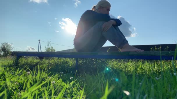 Triste mujer de ensueño sentada en un banco en un pueblo, en el campo. Día soleado de verano, follaje verde por todas partes y rayos de sol brillando en una cámara — Vídeos de Stock