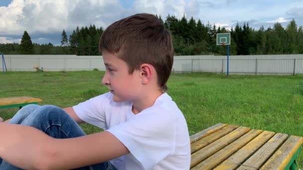 Triste niño soñador pensativo sentado en un banco en un campo de deportes de baloncesto pueblo, campo. Está pensando en la amistad. Día soleado de verano, follaje verde por todas partes — Vídeos de Stock