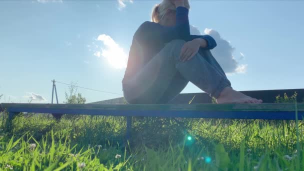 Triste mujer de ensueño sentada en un banco en un pueblo, en el campo. Día soleado de verano, follaje verde por todas partes y rayos de sol brillando en una cámara — Vídeos de Stock