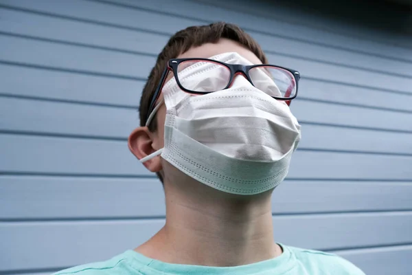 Μια αστεία άποψη ενός αγοριού που φοράει λευκή προστατευτική χειρουργική ιατρική μάσκα, καλύπτει ακόμη και τα μάτια του για την πρόληψη της μόλυνσης κατά τη διάρκεια επιδημιών, γυαλιά του πάνω από τη μάσκα, πανδημία φάρσα rofl αστείο — Φωτογραφία Αρχείου