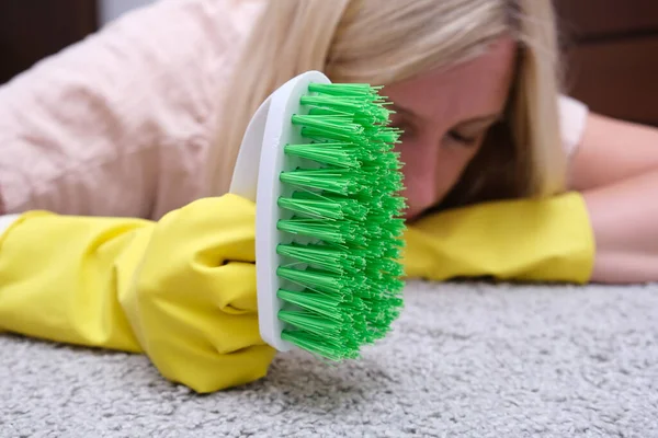 Woman in yellow gloves with a green brush is so tired of cleaning and brushing carpet, removing stains and wool from it and doing routine homework, that she lay down on the floor and sleep