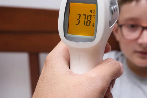 Non-contact infrared thermometer showing high body temperature, unheakthy chuld concept. Mother measuring her son child schoolboy temp at home to make decision about going to school.