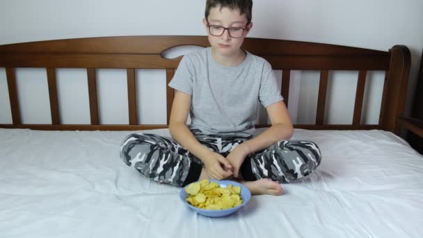 Um menino sentado em um sofá em casa e comendo batatas fritas crocantes de uma tigela — Vídeo de Stock