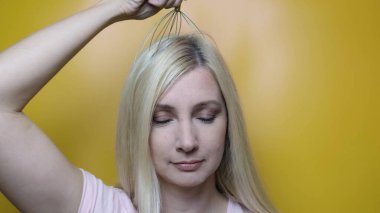 A caucasian blond woman enjoy head scalp massage by anti stress acupuncture metal octopus tool, equipment, she closes her eyes with pleasure, yellow background. clipart