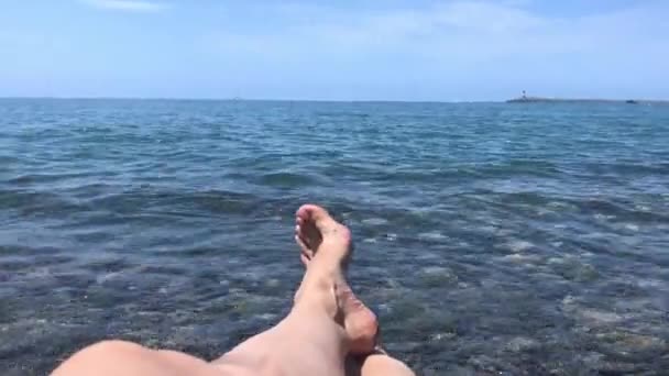 Ноги жінки, що лежать на пляжі і дивляться на морський горизонт з човнами і кораблями і маяком, вода котиться над її ногами — стокове відео
