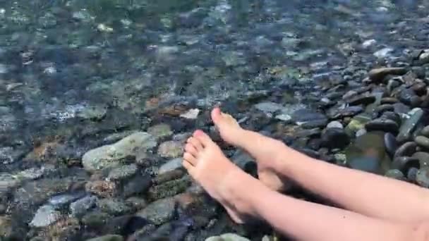Picioarele și picioarele unui copil care stă pe un țărm de mare cu pietricele și se relaxează, apa sărată se rostogolește peste picioare — Videoclip de stoc